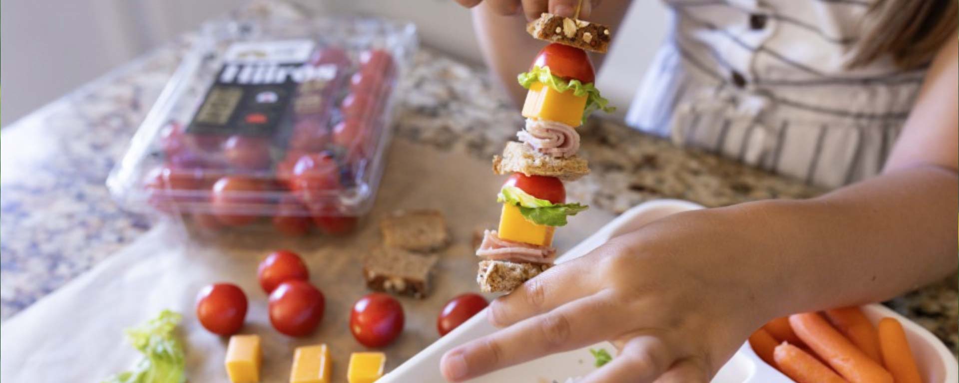 A child assembling a healthy snack skewer with tomatoes, cheese, lettuce, and bread next to a package of Nature Fresh Farms tomatoes.