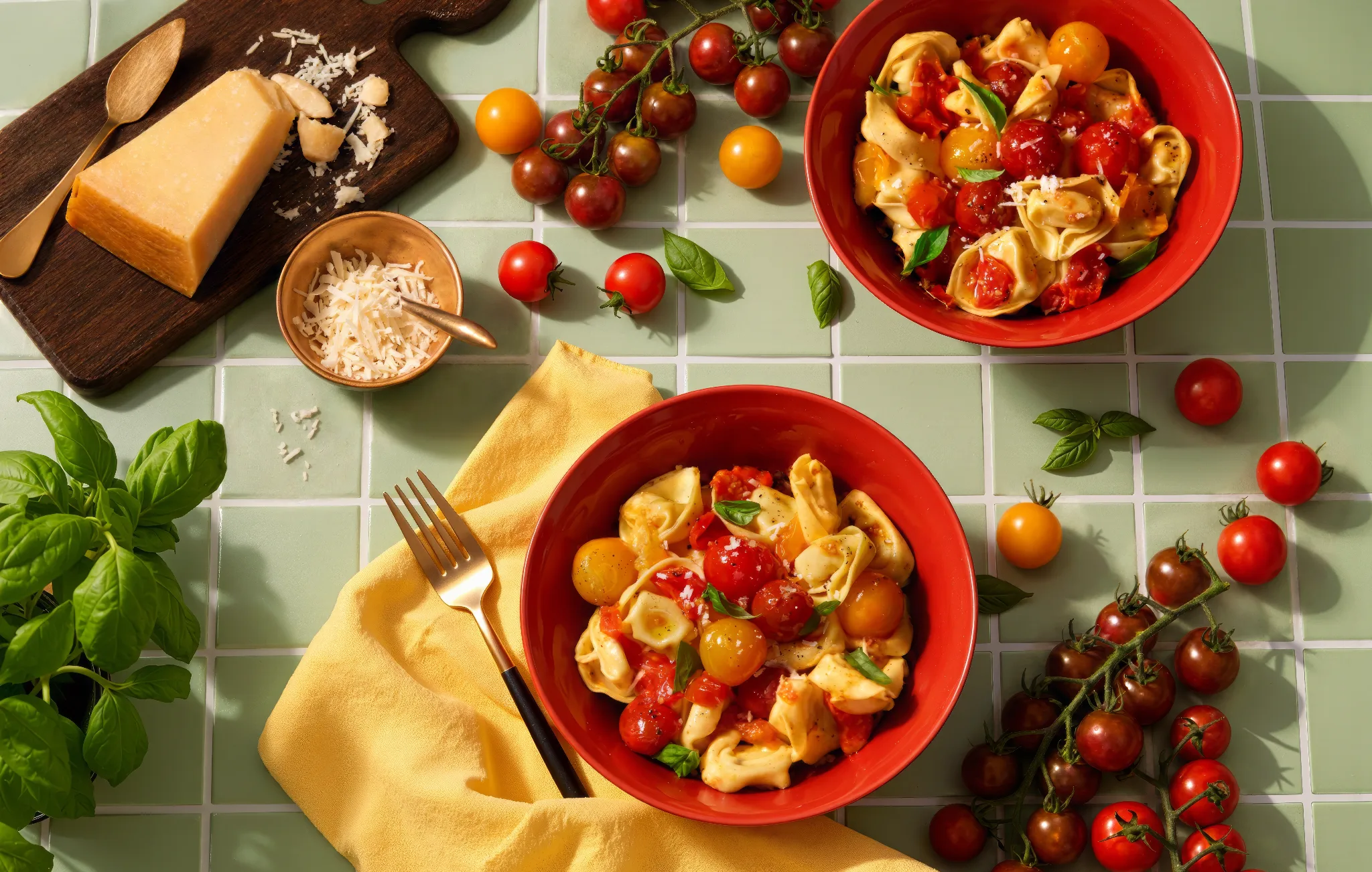 Ombrés™ Tortellini with Garlic Butter in two red bowls, with Ombrés™ tomatoes on the vine, basil and Parmesan